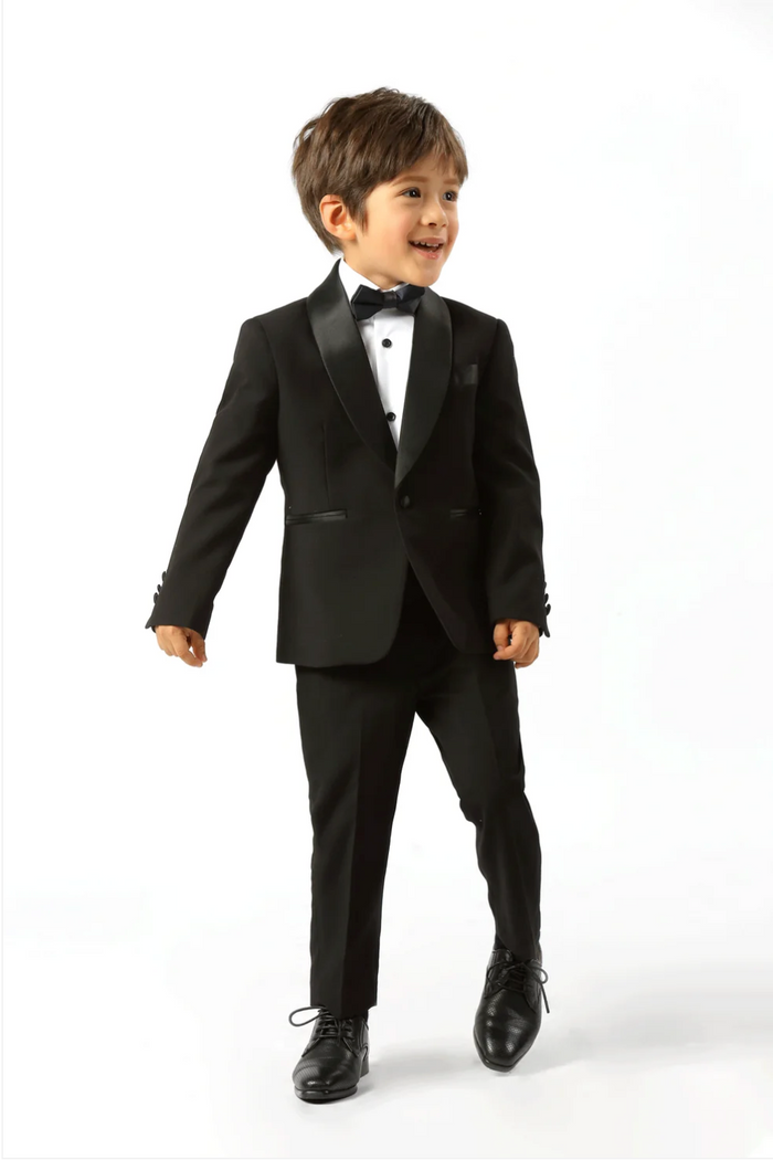A young boy modeling a black kids' tuxedo with satin lapels, a white dress shirt, a black bow tie, and black dress shoes, photographed against a seamless white studio background.