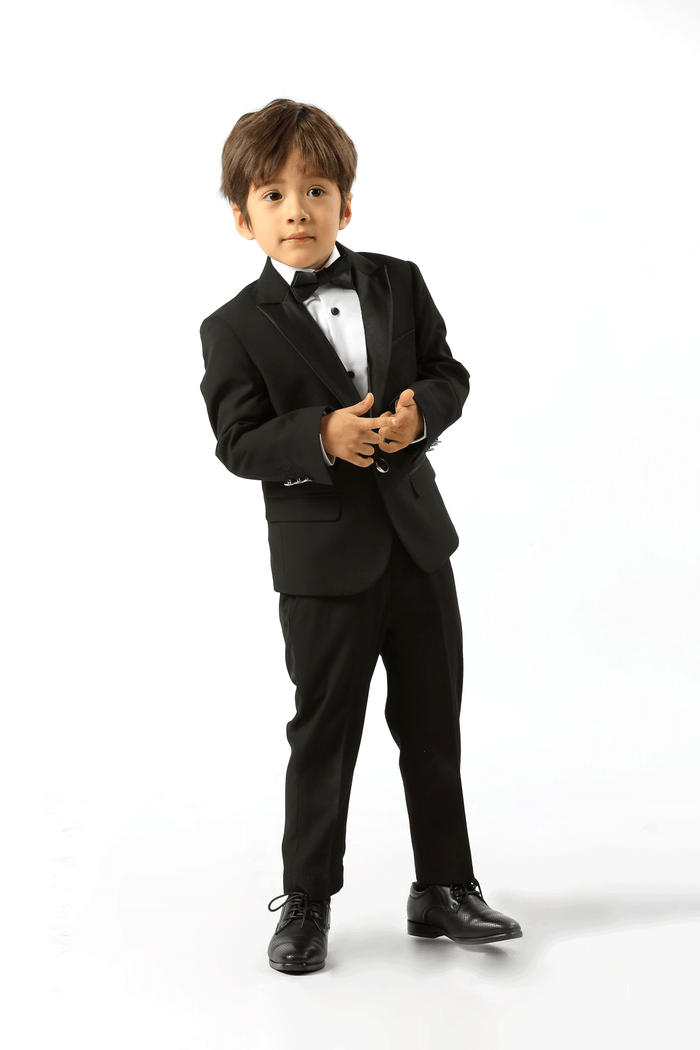 A young boy modeling a black formal tuxedo suit with a white dress shirt, black bow tie, and black dress shoes, standing against a solid white studio background.