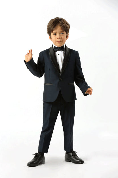 A young boy modeling a dark navy blue kids' tuxedo with black satin lapels, a white dress shirt, and a black bow tie, standing against a plain white studio background.